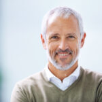 Gray-haired man with dentures smiles while wearing a green sweater