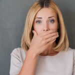 Blonde woman with brown roots covers her mouth in embarrassment because she has bad breath