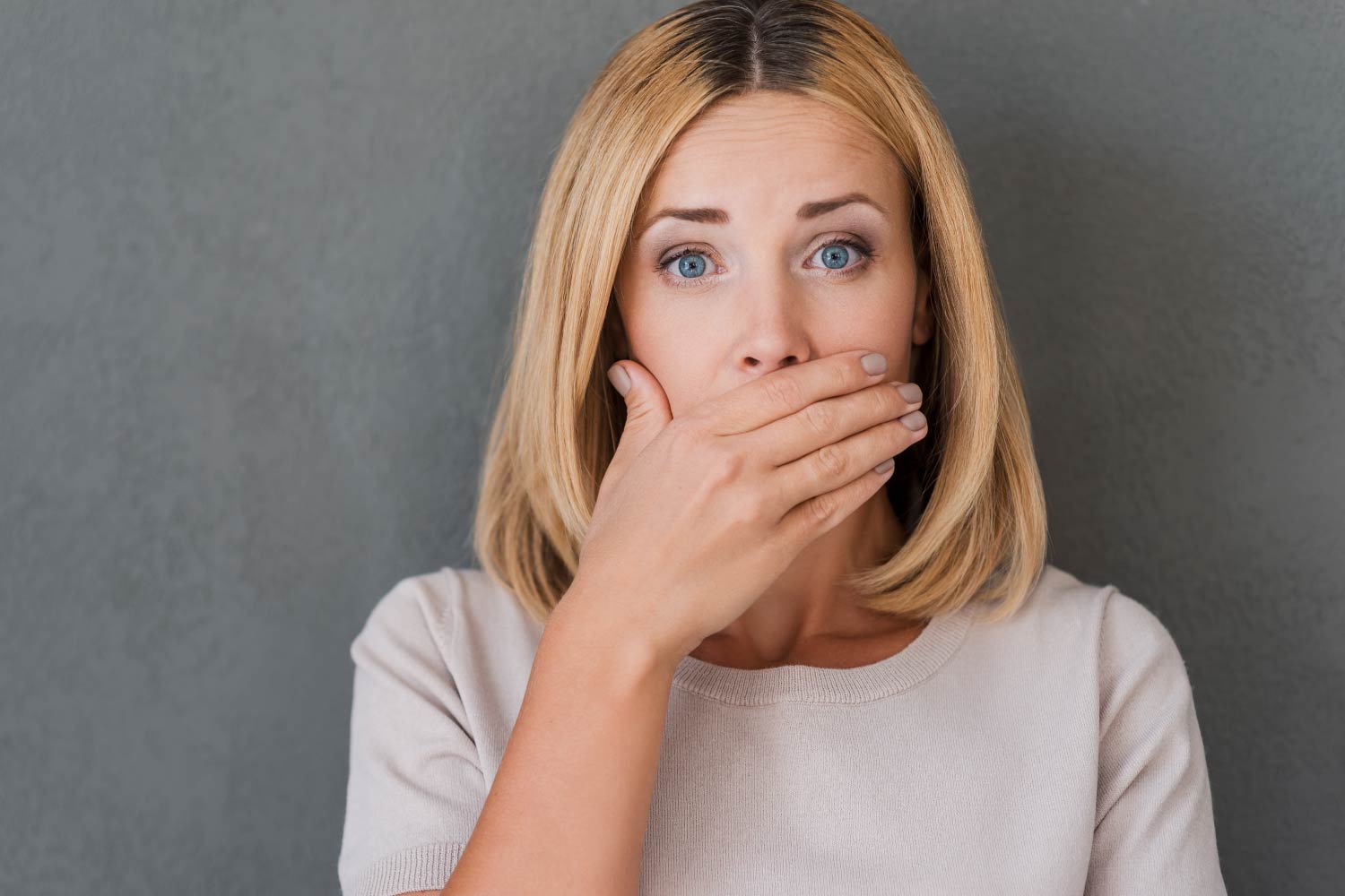 Blonde woman with brown roots covers her mouth in embarrassment because she has bad breath