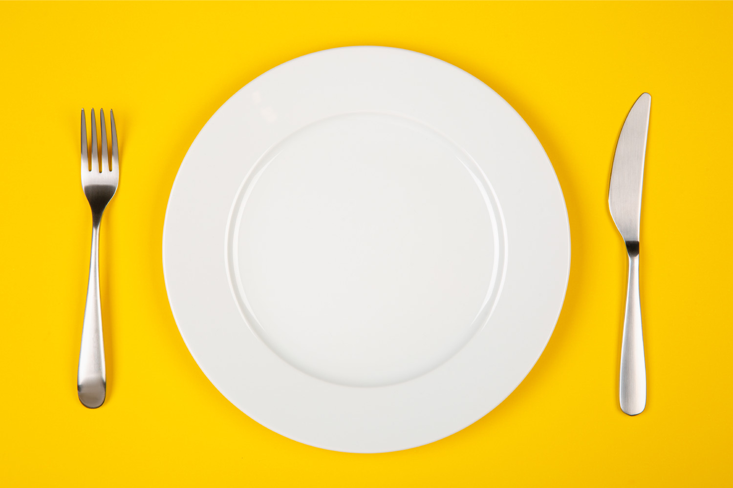 Aerial view of a white plate, fork, and knife on a yellow table for food