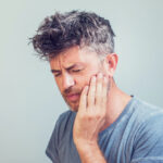Middle-aged white man in a gray shirt cringes in pain and holds his cheek due to a toothache