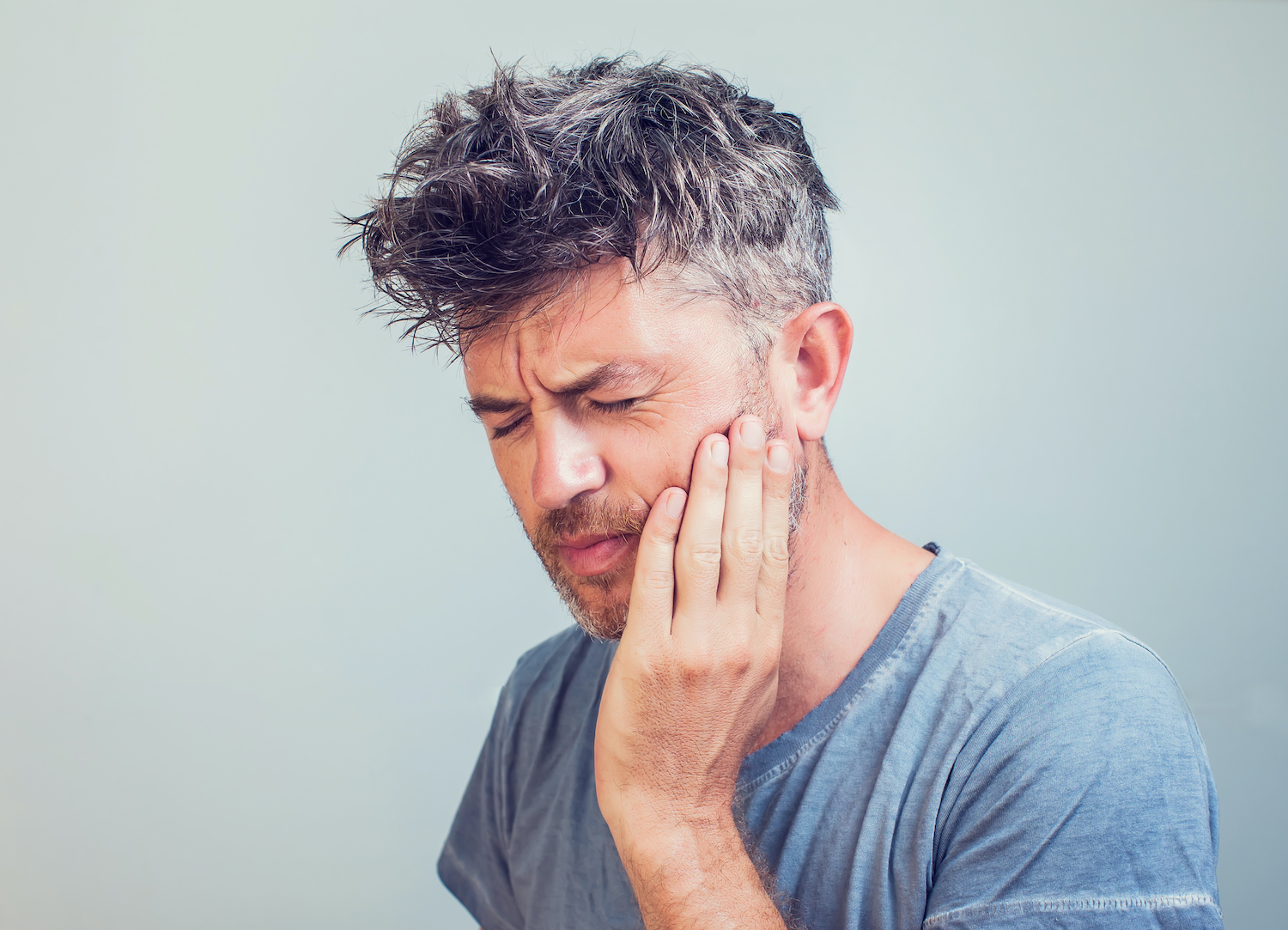 Middle-aged white man in a gray shirt cringes in pain and holds his cheek due to a toothache